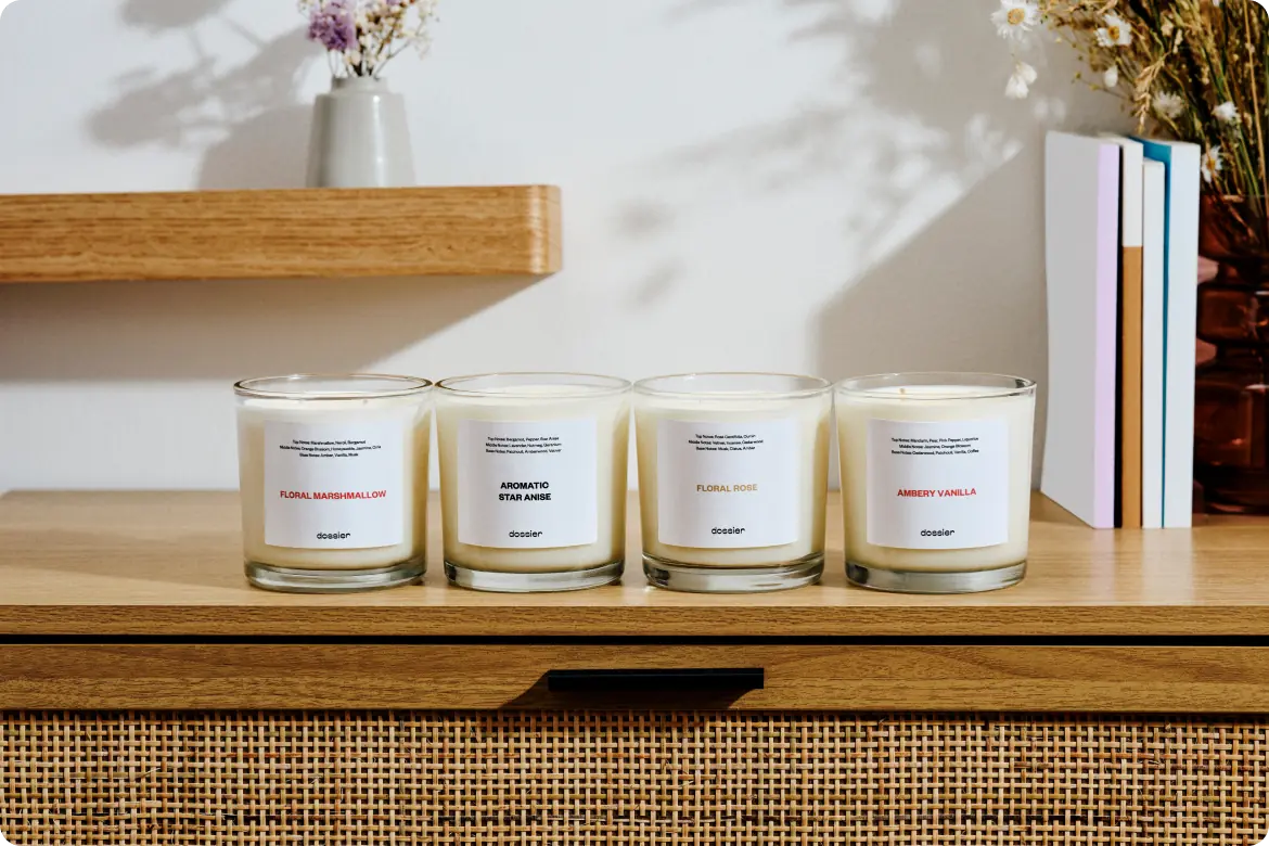 Four Dossier scented candles—Floral Marshmallow, Aromatic Star Anise, Floral Rose, and Ambery Vanilla—displayed on a wooden cabinet in a warm, sunlit interior.