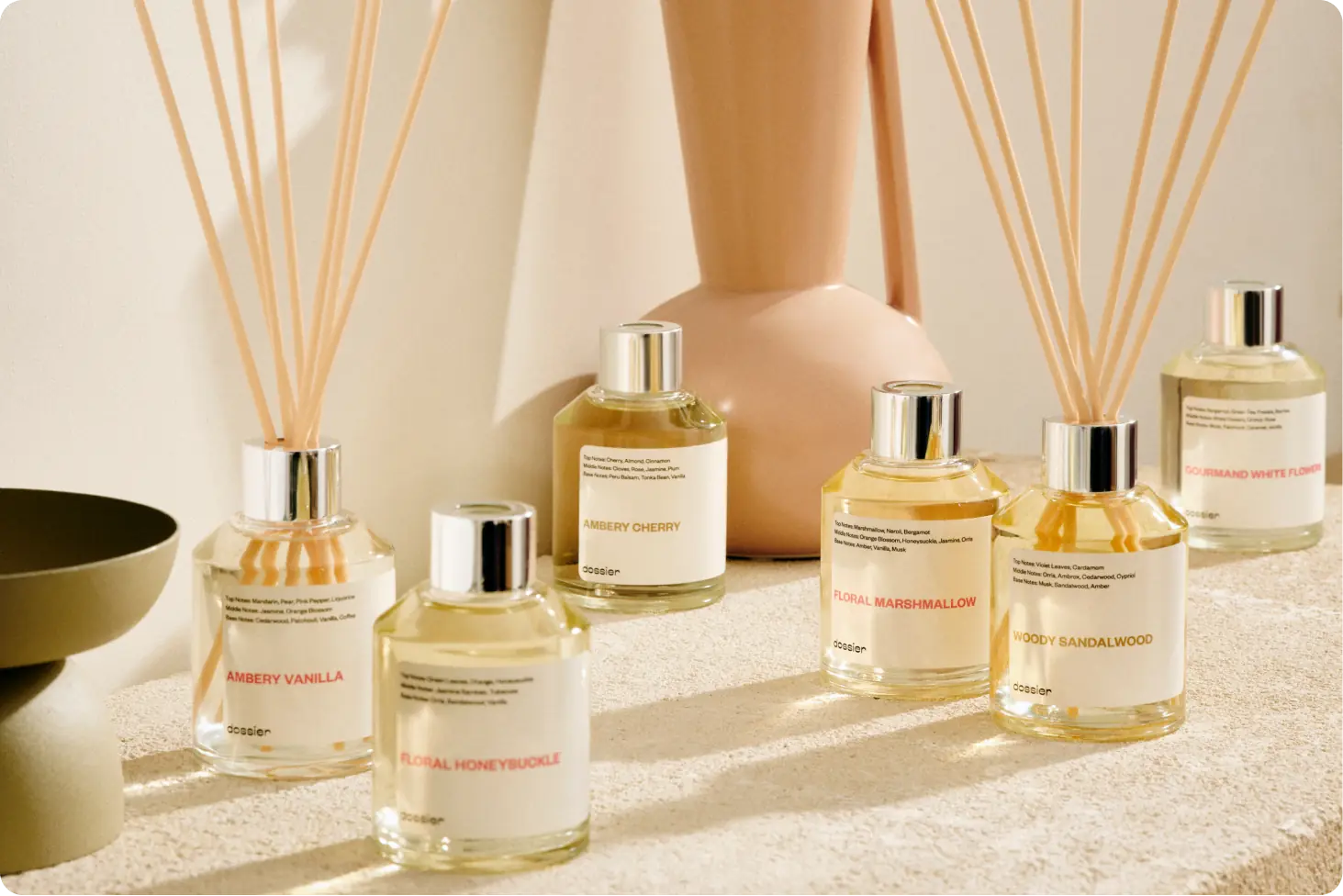 Assorted Dossier reed diffusers, including Ambery Vanilla, Floral Honeysuckle, Ambery Cherry, Floral Marshmallow, Woody Sandalwood, and Gourmand White Flowers, arranged in a warm neutral interior setting.