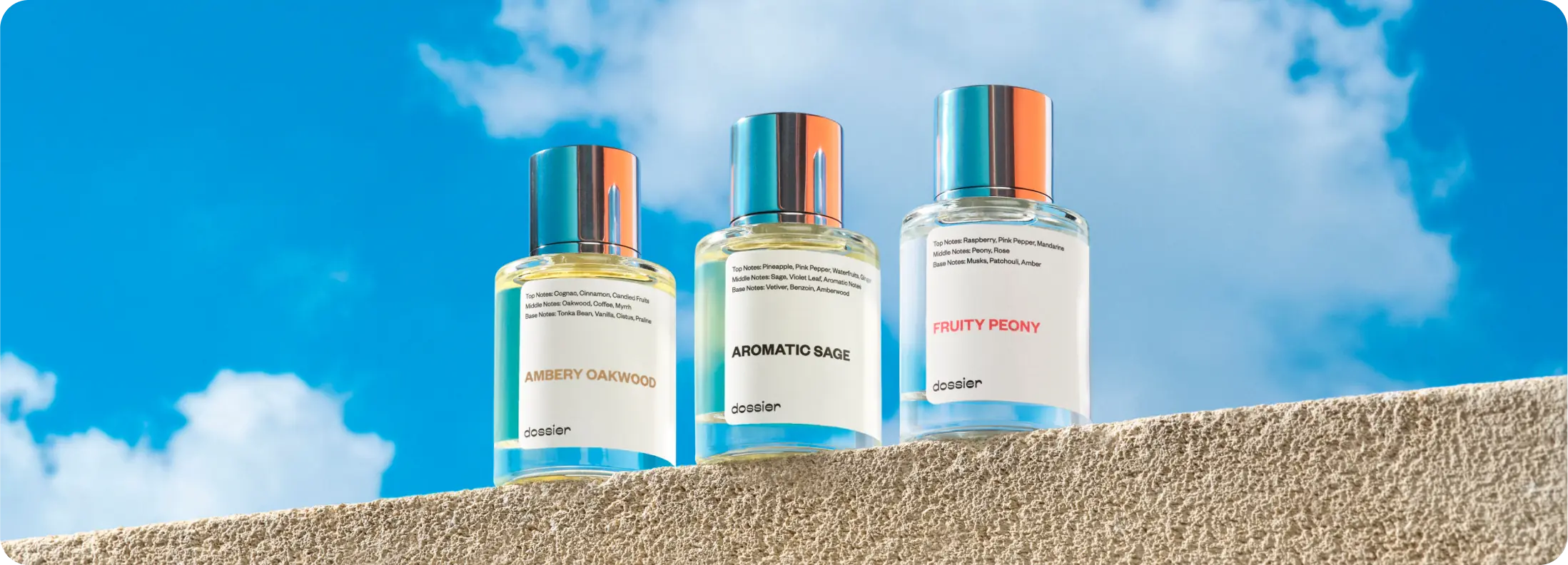 Three Dossier perfume bottles—Ambery Oakwood, Aromatic Sage, and Fruity Peony—displayed on a textured ledge against a bright blue sky with clouds.