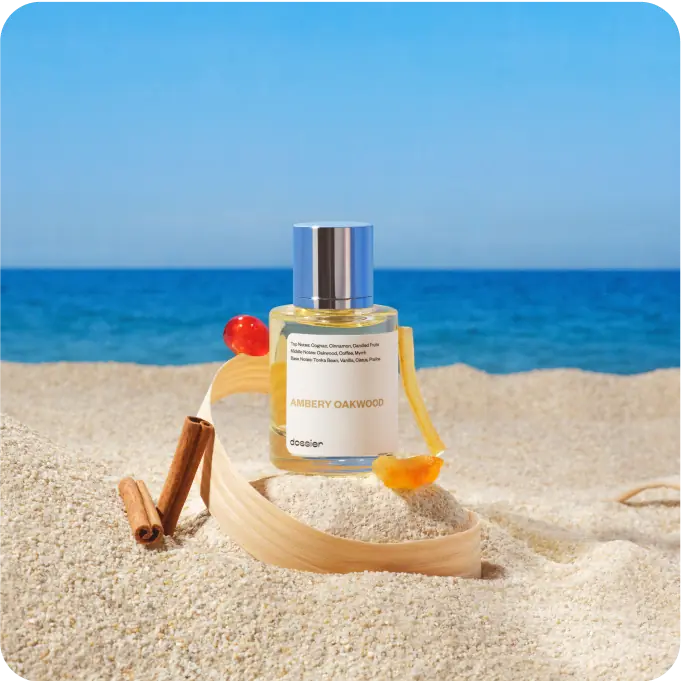 Dossier Ambery Oakwood perfume bottle displayed in sand with cinnamon sticks and warm amber accents, with the sea and blue sky in the background.
