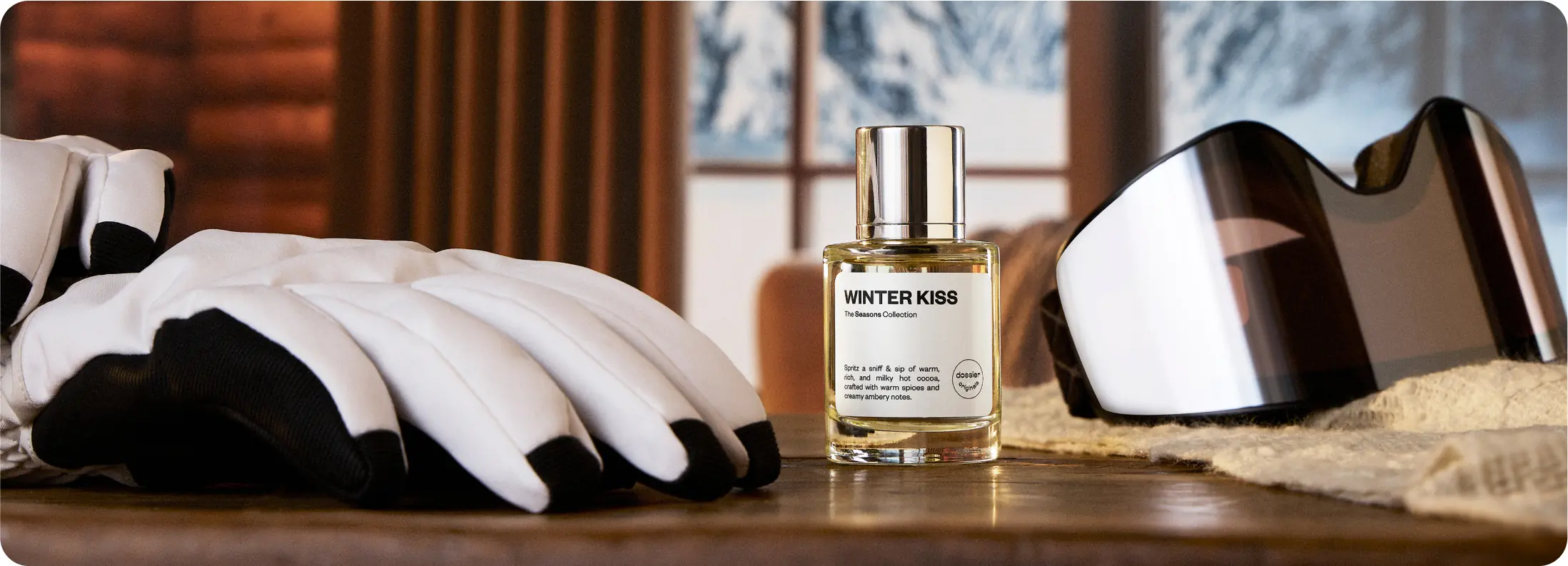 Dossier Winter Kiss perfume bottle displayed beside ski gloves and goggles in a cozy winter setting with snowy mountains visible through the window.