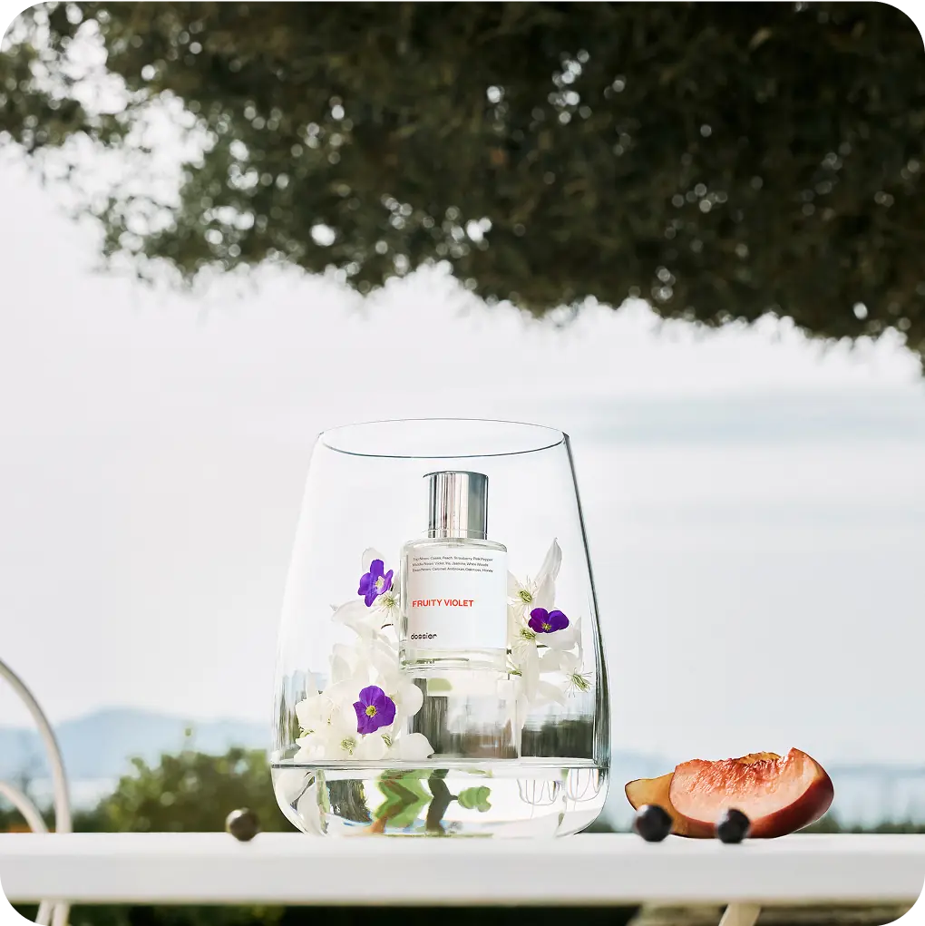 Dossier Fruity Violet perfume displayed in a glass vessel with white and purple flowers, peach, and berries in an outdoor setting.