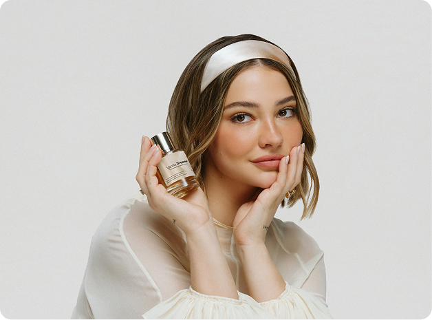 Woman wearing a white headband and cream blouse holding a Vanilla Dreamer perfume bottle beside her face.