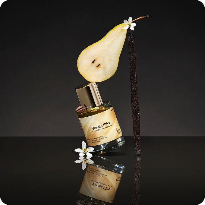 Vanilla Flirt perfume bottle styled with a sliced pear, vanilla pod, coffee bean and delicate white flowers against a dark background.